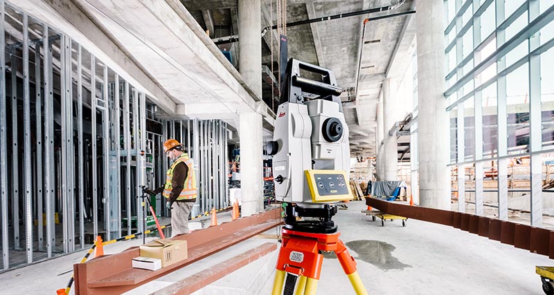Trabalhador da construção com colete laranja e capacete de segurança na obra olha uma controladora Leica iCON CC80 conectada a uma estação total robótica de construção Leica iCON iCR70.